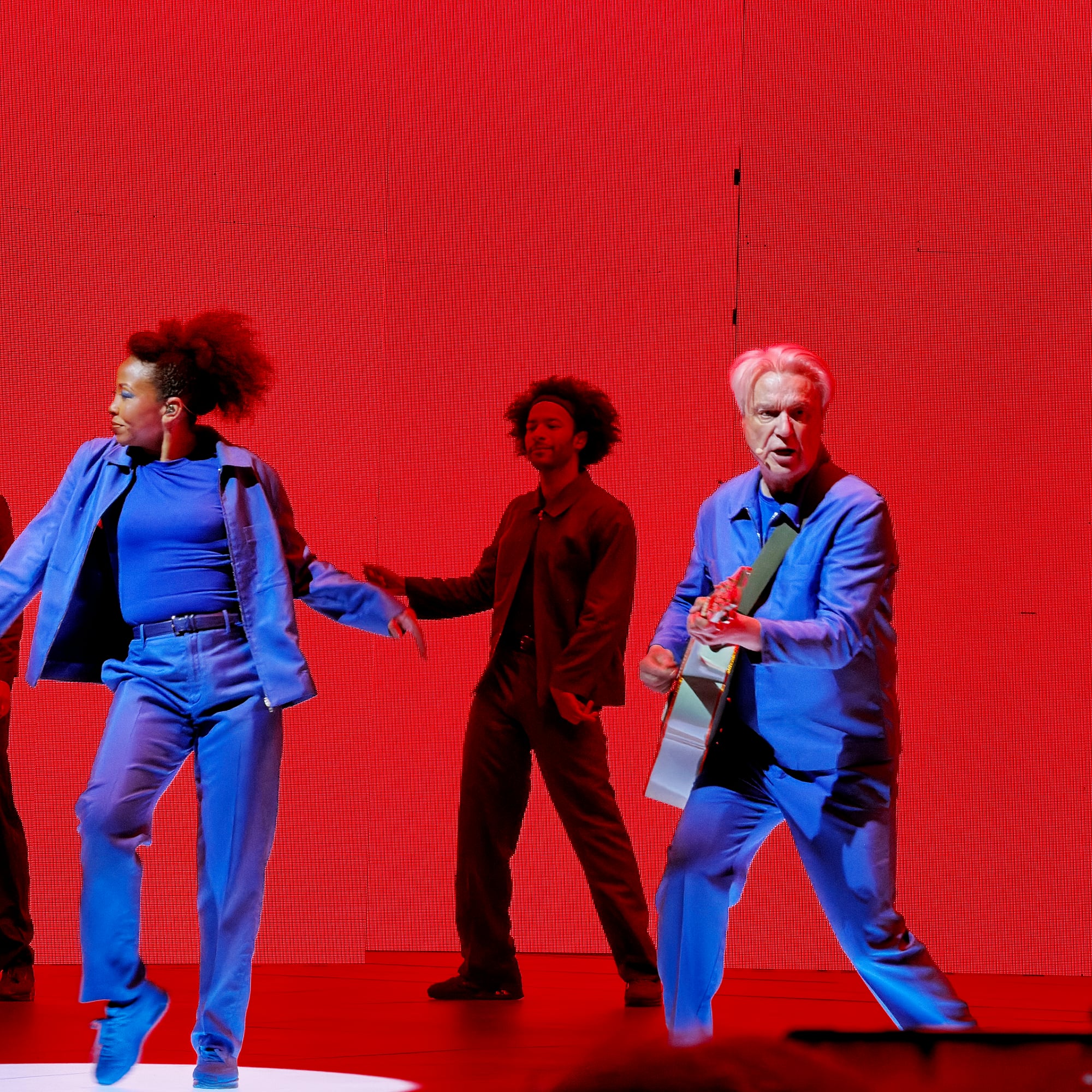 David Byrne playing guitar alongside two dancers, everyone dressed in blue, against a red backdrop